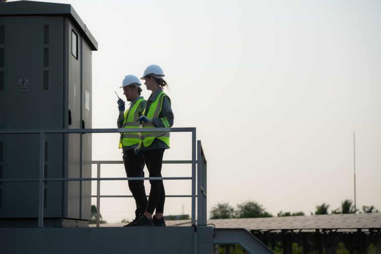 Two workers in hard hats and safety vests on a platform beside electrical equipment, one using a radio