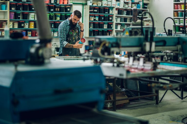 A print shop owner taking a call among shelves of ink and screen printing presses